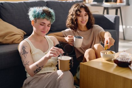 Téléchargez les photos : Jeune amputée avec une main myoélectrique tenant une tasse avec du thé tout en étant assise sur le sol à côté de sa petite amie prenant un cookie dans un bol - en image libre de droit