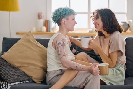 Foto de Mujer joven feliz con taza de té mirando a su novia con discapacidad mientras ambos están sentados en un cómodo sofá delante de la cámara - Imagen libre de derechos