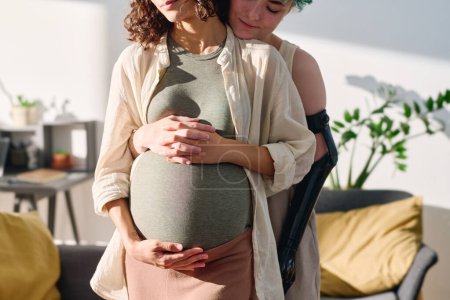 Photo for Cropped shot of sunlit young lesbian couple standing in embrace while girl with myoelectric hand standing behind her pregnant girlfriend - Royalty Free Image