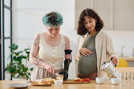 Téléchargez les photos : Jeune femme enceinte verser du café dans une tasse tandis que la fille avec handicap couper baguette fraîche pour sandwichs et préparer le petit déjeuner - en image libre de droit