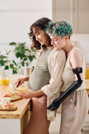 Téléchargez les photos : Jeune femme handicapée embrassant sa petite amie enceinte étalant du beurre sur des tranches de baguette tout en restant debout près de la table de cuisine - en image libre de droit