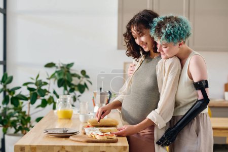 Téléchargez les photos : Vue latérale d'une fille affectueuse avec un handicap embrassant sa petite amie enceinte souriante faisant des sandwichs pour le petit déjeuner dans la cuisine - en image libre de droit