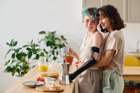 Photo for Young affectionate woman embracing her girlfriend with myoelectric arm holding cup of coffee while standing in front of kitchen table with snacks - Royalty Free Image