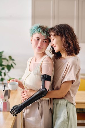 Téléchargez les photos : Jeune femme handicapée tenant une tasse de café et regardant sa petite amie heureuse tout en restant debout près de la table de cuisine - en image libre de droit