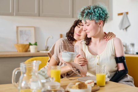 Foto de Joven morena abrazando a su novia con brazo mioeléctrico tomando té o café mientras ambos están sentados junto a la mesa servida y desayunando - Imagen libre de derechos