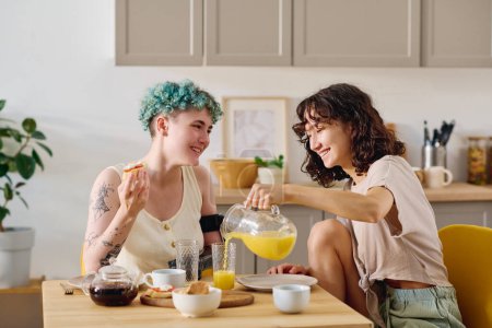 Photo for Young smiling brunette woman pouring fresh orange juice into glass of her girlfriend with disability eating sandwich and chatting to her by breakfast - Royalty Free Image