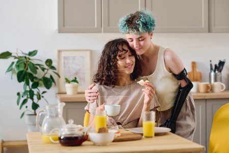 Téléchargez les photos : Jeune femme affectueuse avec la main myoélectrique donnant câlin à jolie petite amie brune ayant une tasse de café et sandwich fait maison - en image libre de droit