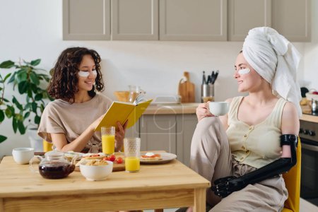 Foto de Dos lindas novias sonrientes con parches debajo de los ojos sentadas junto a la mesa de la cocina servidas con aperitivos y bebidas, charlando y leyendo el libro para el desayuno - Imagen libre de derechos