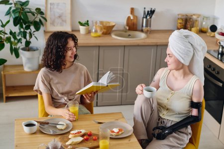 Téléchargez les photos : Jeune femme brune souriante avec livre ouvert regardant sa petite amie handicapée assise près d'une table de cuisine servie et bavardant au petit déjeuner - en image libre de droit