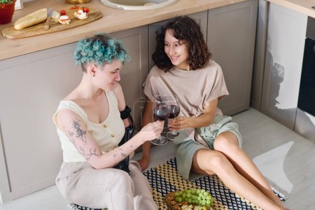Photo for Happy young brunette woman and her girlfriend with disability clinking with glasses of red wine while sitting on the floor by kitchen counter - Royalty Free Image