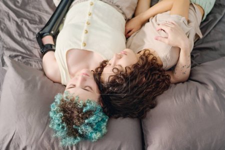 Téléchargez les photos : Au-dessus de l'angle de la jeune femme handicapée embrassant sa jolie petite amie endormie tout en étant couchée sur un lit double confortable et en faisant la sieste - en image libre de droit
