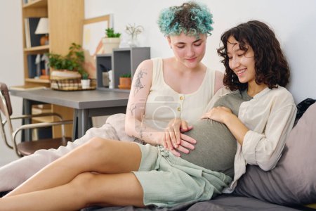 Photo for Young woman with myoelectric hand touching belly of smiling pregnant girlfriend while both sitting on double bed and spending time together - Royalty Free Image