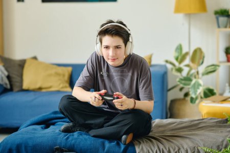 Foto de Morena adolescente en auriculares presionando botones en el mando mientras está sentado en la cama delante de la computadora y jugando videojuego en casa - Imagen libre de derechos