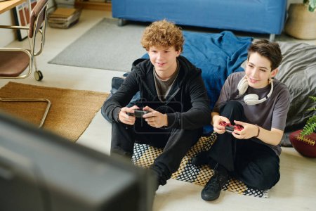 Téléchargez les photos : Deux adolescents souriants passer leurs loisirs à la maison et jouer à des jeux vidéo tout en étant assis sur le sol et en appuyant sur les boutons sur les manettes - en image libre de droit