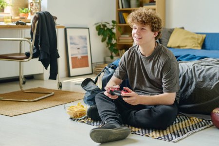 Foto de Adolescente con el pelo rizado sentado en el suelo por la cama y jugando a videojuegos mientras mira la pantalla de la computadora y presiona los botones de la consola - Imagen libre de derechos