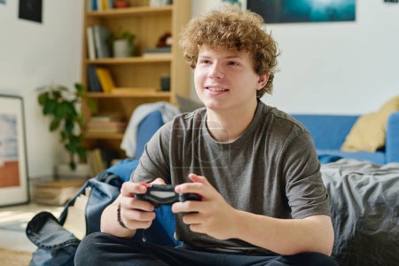 Téléchargez les photos : Adolescent souriant mignon avec joystick appuyant sur les boutons pendant le jeu d'ordinateur tout en étant assis au lit devant la caméra et profiter de loisirs - en image libre de droit