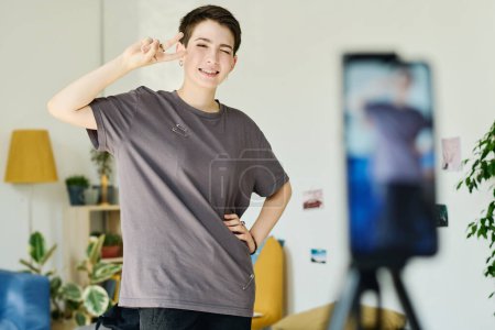 Photo for Happy girl in grey t-shirt showing peace gesture and looking at smartphone camera while shooting livestream for online audience - Royalty Free Image