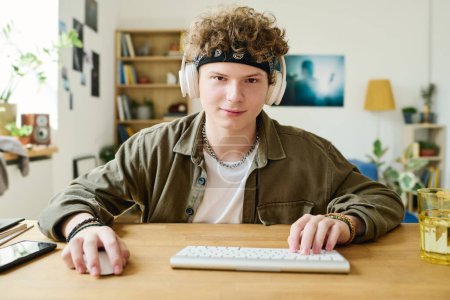 Téléchargez les photos : Adolescent mignon gardant les mains sur la souris et les boutons du clavier tout en étant assis par le bureau devant l'ordinateur et en regardant les données à l'écran - en image libre de droit