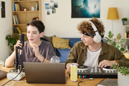 Photo for Teenage guy in headphones looking at his girlfriend talking in microphone while sitting next to her and adjusting turntables during livestream - Royalty Free Image