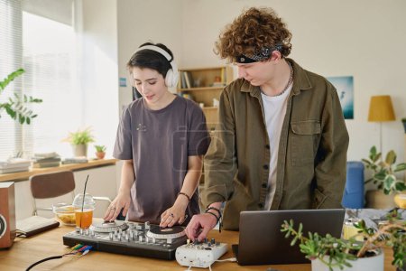 Téléchargez les photos : Couple adolescent moderne créant une nouvelle musique informatique et l'enregistrant tout en restant debout au bureau avec des platines tournantes et un ordinateur portable dans l'environnement de la maison - en image libre de droit