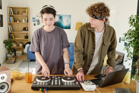 Téléchargez les photos : Adolescent gars en tenue décontractée regardant sa petite amie dans les écouteurs ajuster les platines tournantes tout en créant de la nouvelle musique dans l'environnement de la maison - en image libre de droit