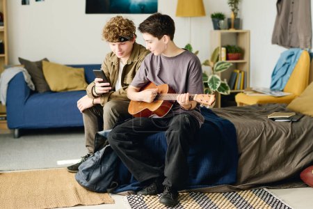 Photo for Long shot of two teenagers in casualwear looking at smartphone screen while sitting on bed at home and shooting video for online audience - Royalty Free Image