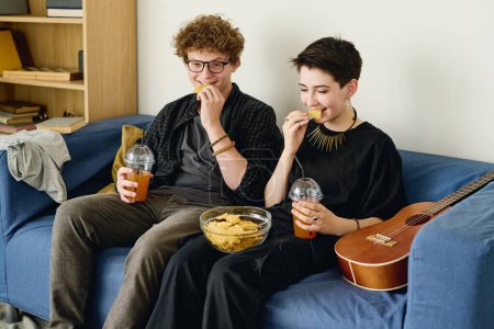 Téléchargez les photos : Adolescent affamé gars et fille manger des croustilles de bol en verre et prendre des boissons tout en étant assis sur le canapé dans le salon à loisir - en image libre de droit