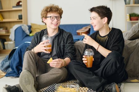 Téléchargez les photos : Adolescente heureuse avec un verre de jus ou de soda regardant son petit ami tandis que les deux assis sur le sol dans la chambre, avoir une collation et bavarder - en image libre de droit