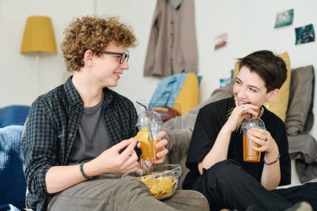 Téléchargez les photos : Deux adolescents heureux mignons prendre des boissons, croustilles et discuter de quelque chose tout en étant assis au lit et profiter des loisirs ou week-end - en image libre de droit