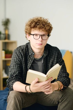 Photo for Serious teenage boy in eyeglasses with open book of stories or novel looking at camera while sitting on bed and spending leisure at home - Royalty Free Image