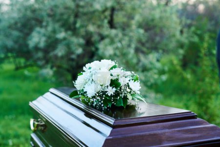 Téléchargez les photos : Cercueil en bois recouvert d'un lourd couvercle avec un bouquet de fleurs blanches debout contre des arbres verts et des buissons sur le cimetière avant l'enterrement - en image libre de droit