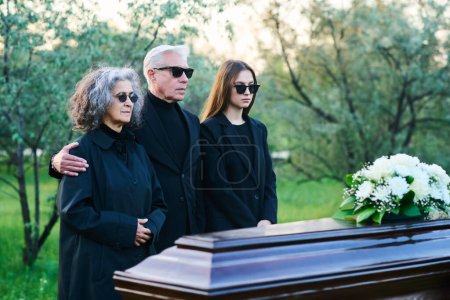 Téléchargez les photos : Mature mari et femme et leur fille en noir total debout par cercueil avec un bouquet de fleurs blanches sur sa couverture tout en enterrant leur parent - en image libre de droit