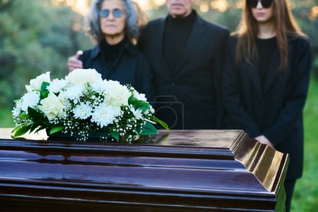 Bande de roses blanches et de chrysanthèmes sur le dessus du cercueil fermé debout devant la caméra contre les personnes portant des vêtements de deuil