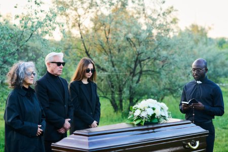 Téléchargez les photos : Pasteur afro-américain avec Bible ouverte et famille en deuil en tenue noire et lunettes de soleil debout près du cercueil couvert au service funéraire - en image libre de droit