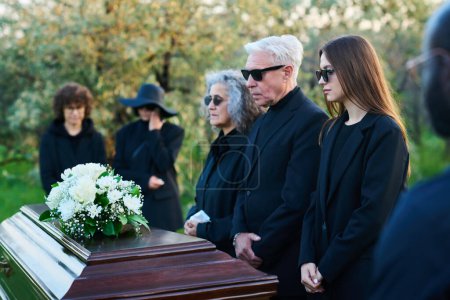 Foto de Familia de tres personas afligidas en gafas de sol y ropa de luto de pie frente al ataúd con flores blancas en la tapa durante el servicio fúnebre - Imagen libre de derechos