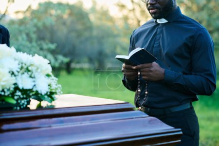 Cropped shot of young priest in black clothes reading verses from Holy Bible during funeral service while standing in front of coffin with flowers
