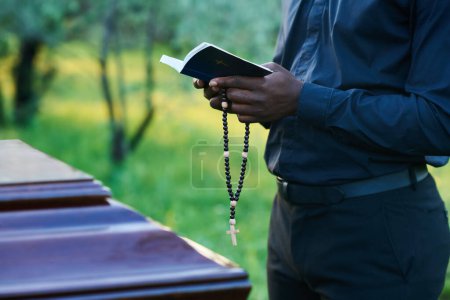 Photo for Close-up of African American man in priest attire holding rosary beads and open Holy Bible while standing in front of coffin during burial service - Royalty Free Image