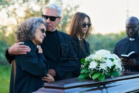 Foto de Familia de luto de tres y sacerdote en traje negro de pie frente al ataúd con flores blancas en la parte superior durante el funeral en el cementerio - Imagen libre de derechos