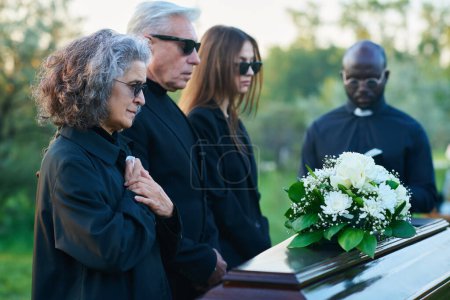 Trauernde reife Frau mit Taschentuch, die ihre Hände auf der Brust hält, während sie während der Trauerfeier vor dem Sarg mit geschlossenem Deckel steht