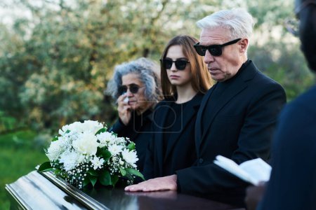 Téléchargez les photos : Homme mûr en lunettes de soleil et tenue de deuil gardant la main sur le couvercle du cercueil fermé tout en se tenant entre sa famille et le prêtre avec la Bible - en image libre de droit