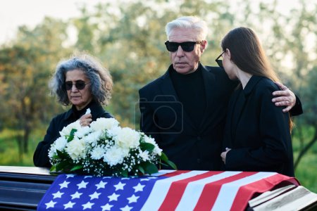 Téléchargez les photos : Homme mûr en costume de deuil embrassant sa fille en deuil tout en se tenant près du cercueil avec des fleurs blanches sur le dessus du couvercle fermé aux funérailles - en image libre de droit