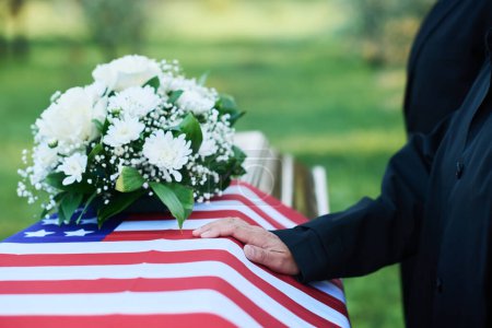 Hand of mature woman in mourning attire on American flag covering coffin with bunch of resh white chrysanthemums and roses on top