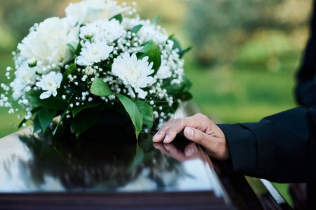 Konzentration auf die Hand einer trauernden reifen Frau in schwarzer Kleidung auf dem Deckel eines geschlossenen Holzsarges mit einem Strauß frischer weißer Chrysanthemen darüber