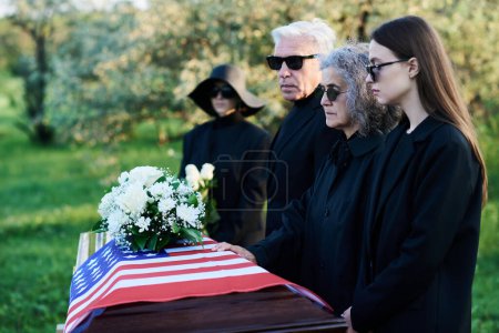 Foto de Fila de familiares con gafas de sol y ropa de luto de pie junto al ataúd cubierto con la bandera estadounidense y lamentando a su pariente muerto - Imagen libre de derechos