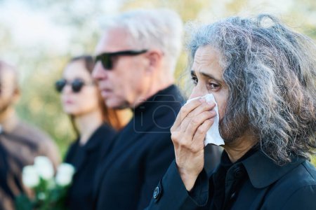 Grieving senior woman wiping tears by handkerchief while crying at funeral of her friend, colleague, relative or family member against other people
