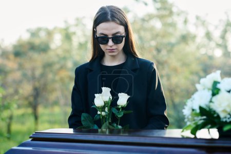 Foto de Joven viuda con racimo de rosas blancas frescas de pie junto al ataúd con la tapa cerrada y flores en su parte superior mientras está de duelo por su marido muerto - Imagen libre de derechos