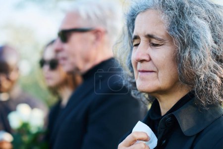 Mature crying woman with tears on her cheek lamenting her relative, family member or friend at funeral service against other people