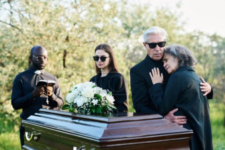 Foto de Hombre maduro apoyando a su esposa inconsolable mientras ambos lloran por su miembro muerto de la familia por ataúd con tapa cerrada en el funeral - Imagen libre de derechos