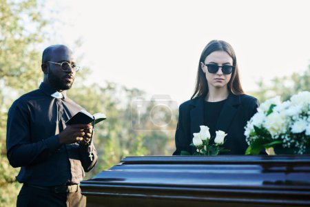 Téléchargez les photos : Jeune veuve inconsolable en costume de deuil et lunettes de soleil tenant un bouquet de roses blanches tout en restant près du cercueil à côté du prêtre avec la Bible - en image libre de droit