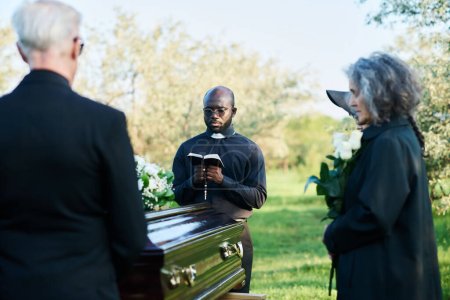 Focus on young African American priest with open Bible standing in front of coffin with closed lid and group of grieving people in black attire
